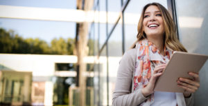 Smiling woman holding a tablet outdoors, wearing a cozy sweater and floral scarf, with modern glass architecture in the background.